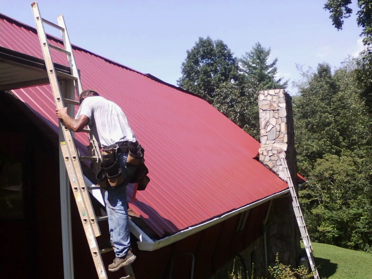 Roofer on a ladder beside a red metal roof and stone chimney — Soffit & Fascia Repair by Roofing Brothers in Mascoutah